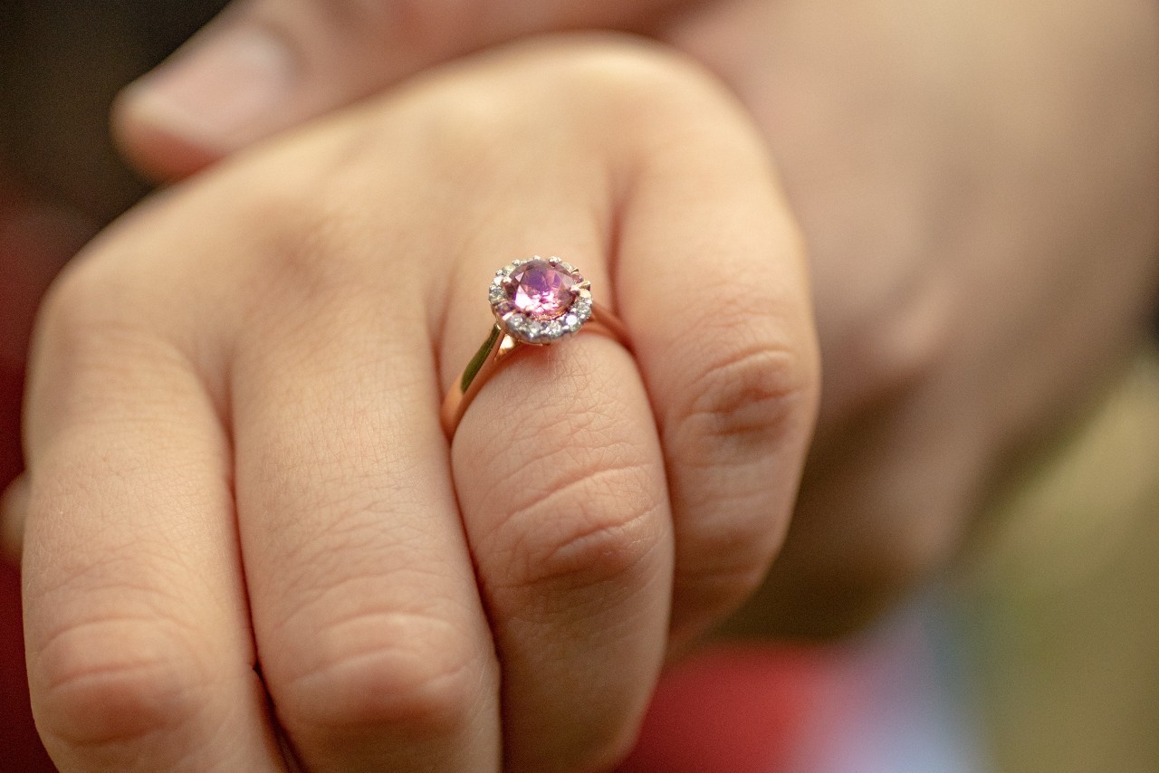 close up of a woman’s hand wearing a rose gold engagement ring with a purple center stone and diamond halo