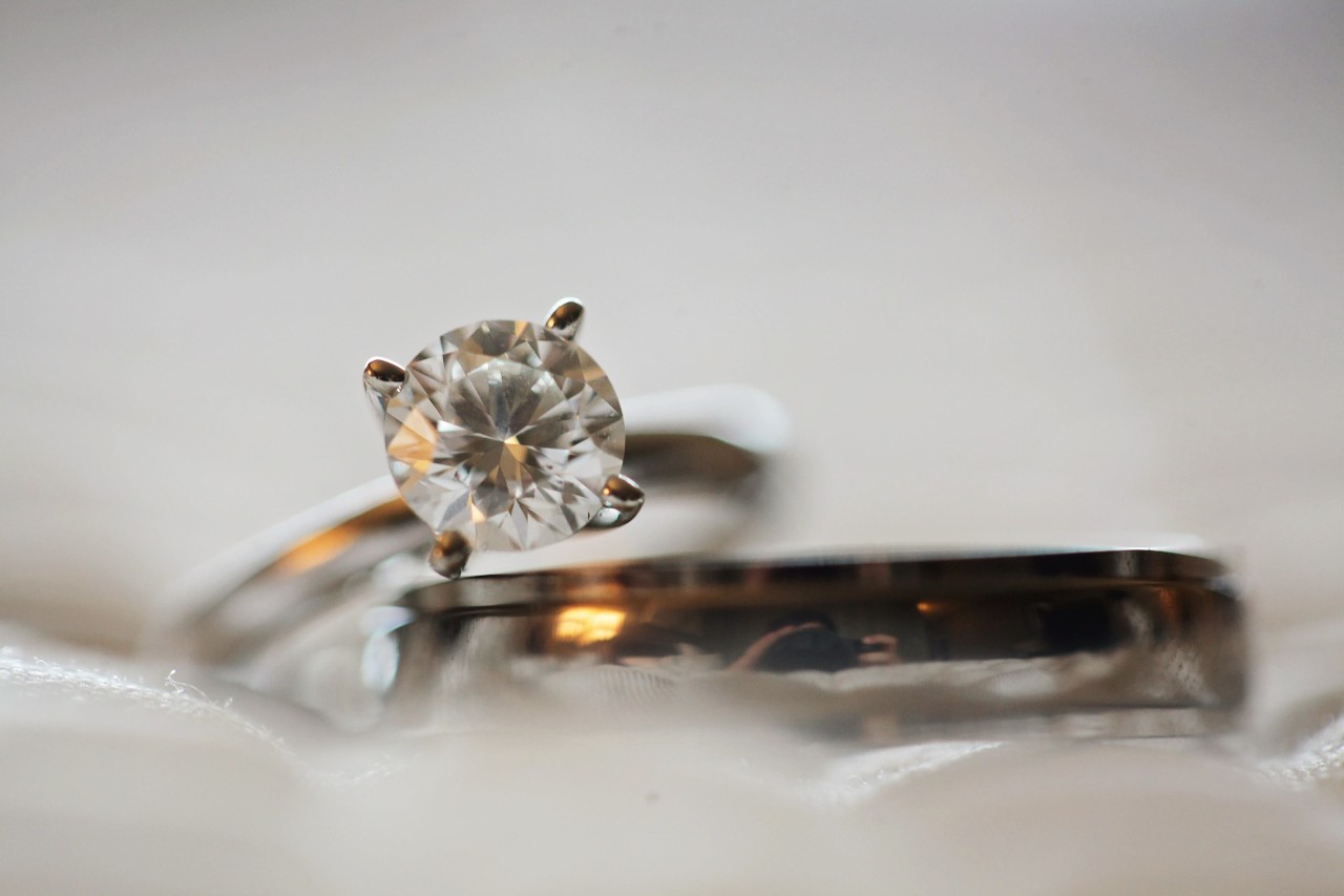 A solitaire engagement ring and a men’s wedding band sit on a white ribbon