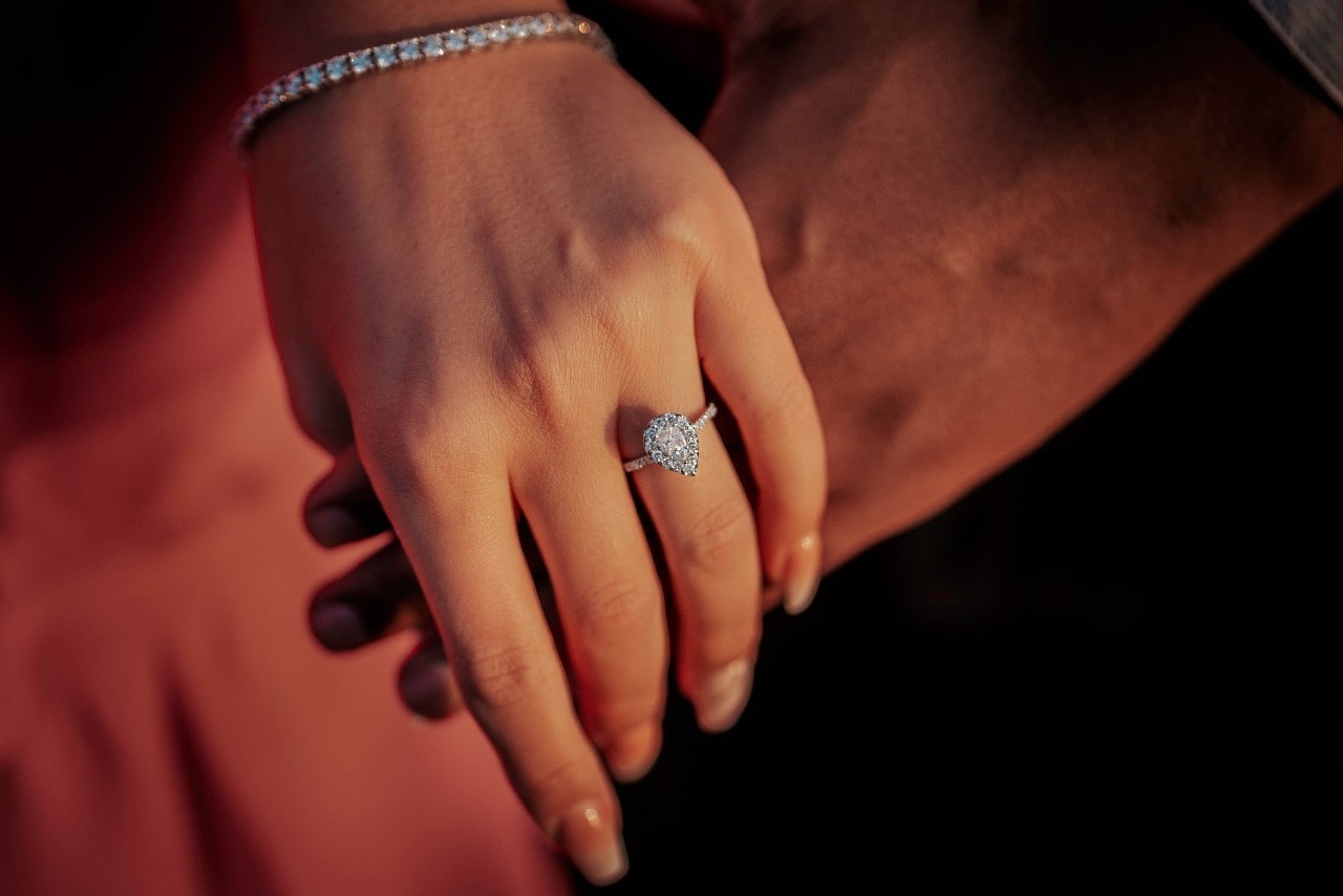 A couple holding hands, the woman wearing a halo engagement ring and a tennis bracelet.